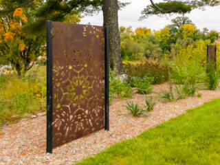 A metal panel with cutouts for a birch bark biting pattern radiating from a center point and symmetrical across four axes, on a stone path with grass, gardens and trees around.