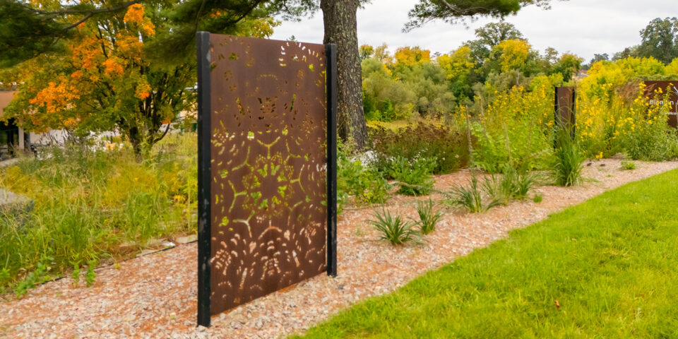 A metal panel with cutouts for a birch bark biting pattern radiating from a center point and symmetrical across four axes, on a stone path with grass, gardens and trees around.